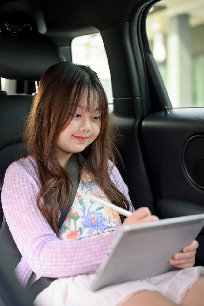 Enfant dessinant sur une tablette en voyage en voiture Enfant souriante dessinant sur une tablette avec un stylet dans une voiture, vêtue d'un gilet lilas et d'une robe fleurie.