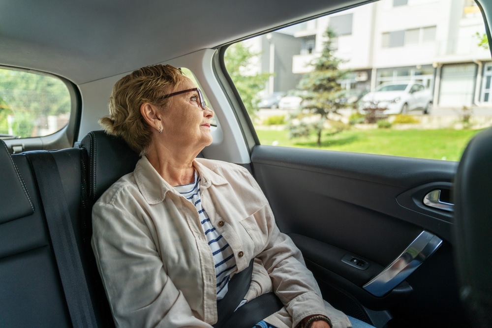 Femme senior pensive en voiture : voyage et sérénité Femme senior pensive regardant par la fenêtre à l'arrière d'une voiture, portant des lunettes et une veste beige.
